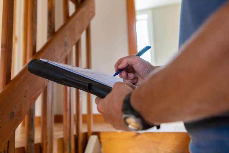 A close up view on the hands of a professional building inspector, standing near a staircase filling in forms on overall environmental quality indoors.